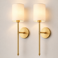 Set of two gold wall sconces with tall cylindrical white shades on a plain background.