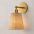 A modern wall-mounted sconce with a warm brass-finished circular backplate, horizontal arm, and cylindrical neck, paired with a tapered woven rattan shade that glows with a warm light against a plain off-white wall.