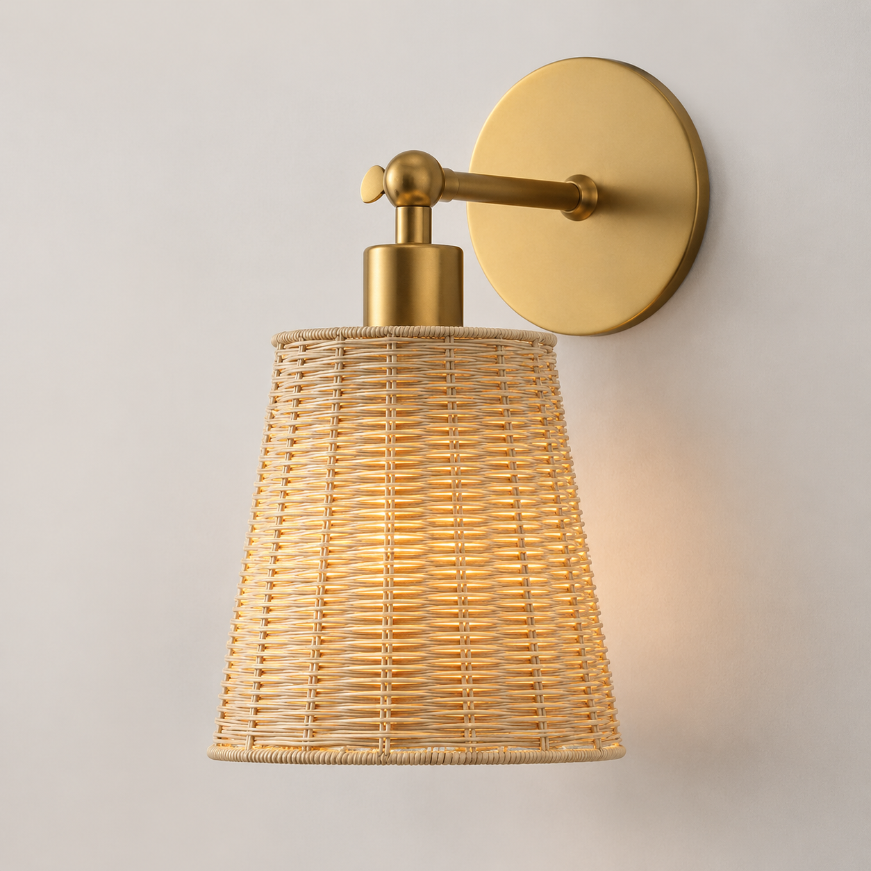 A modern wall-mounted sconce with a warm brass-finished circular backplate, horizontal arm, and cylindrical neck, paired with a tapered woven rattan shade that glows with a warm light against a plain off-white wall.