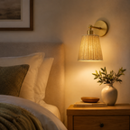 A warm brass wall sconce with a woven natural-fiber shade glowing with a golden light, mounted on a beige wall above a wooden nightstand with a vase of greenery.
