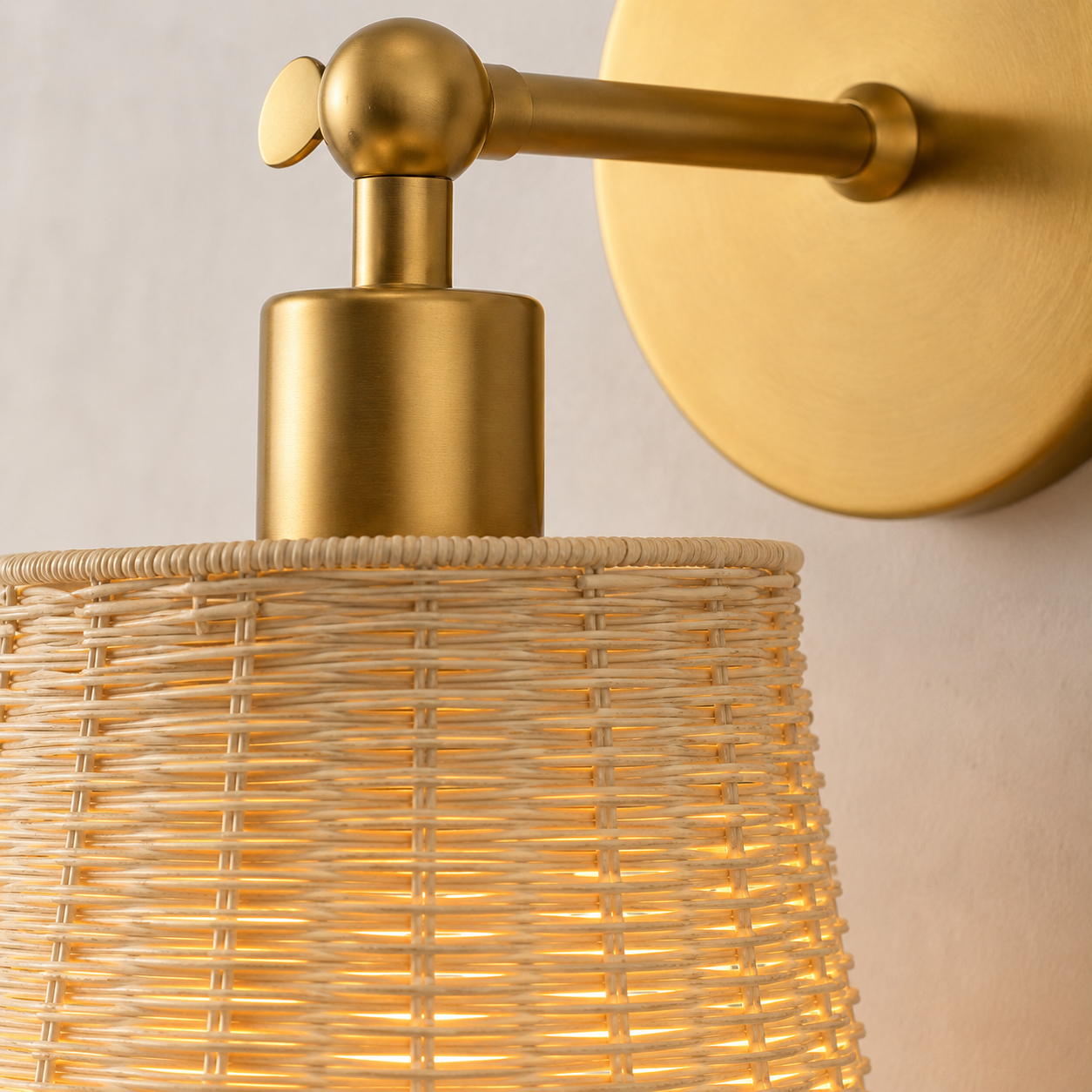 A close-up of a wall-mounted light fixture with a warm brass/gold metal wall plate, arm, and pump-like top, paired with a woven natural-fiber shade that glows with a warm yellow light against a light neutral wall.