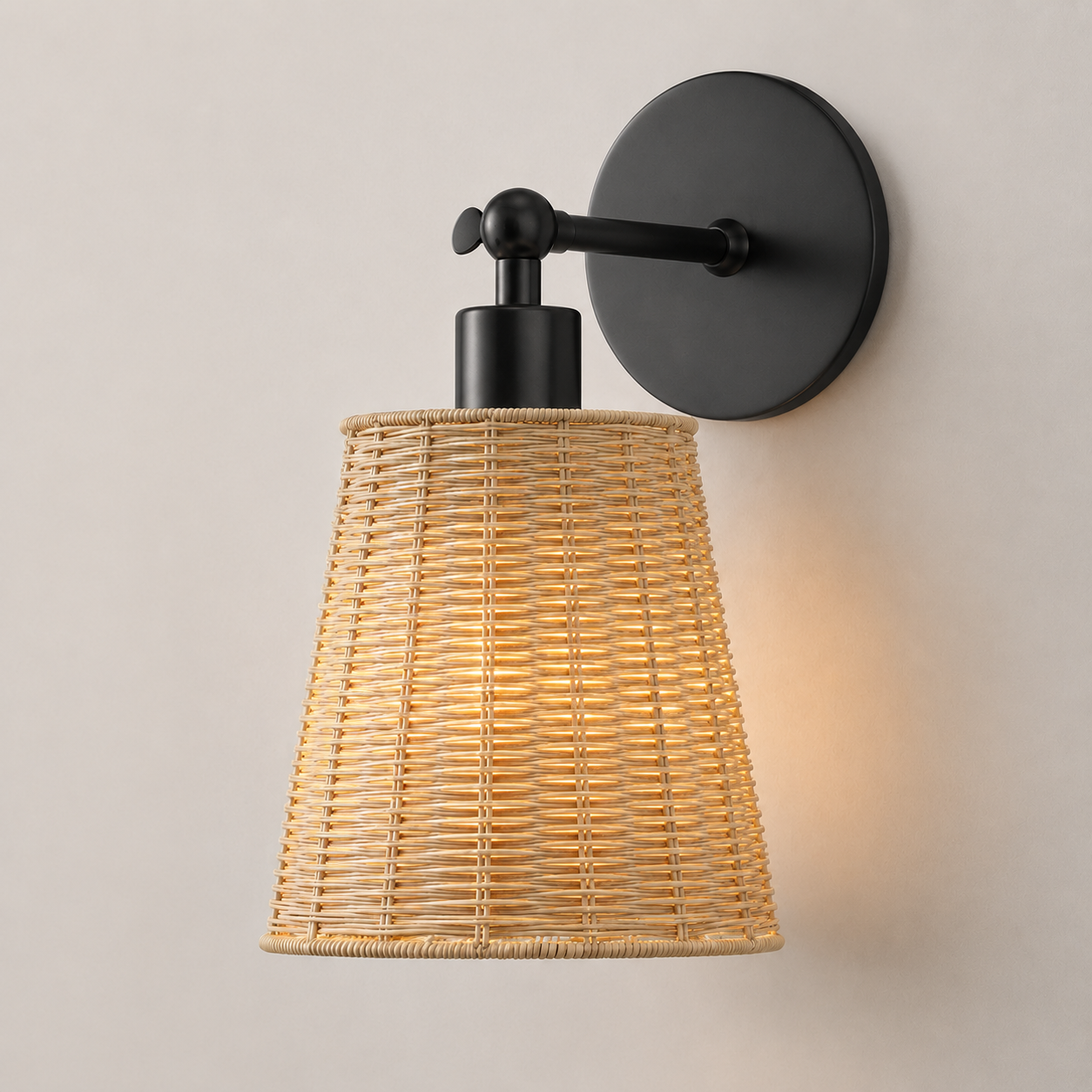 A wall-mounted sconce with a black metal wall plate and arm, featuring a natural woven rattan shade that glows warm yellow when lit, shown against a plain light beige wall.