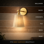 A wall-mounted sconce with a woven beige shade and gold-toned hardware, shown lit against a textured wall with overlaid text listing surfaces like wallpaper, wood, concrete, tile, brick, and paint, plus the phrase 'FITS MANY SURFACES BEAUTIFULLY.'.