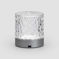 A modern cylindrical table lamp with a textured, hammered clear glass shade and a matte dark gray base, shown on a light gray background.