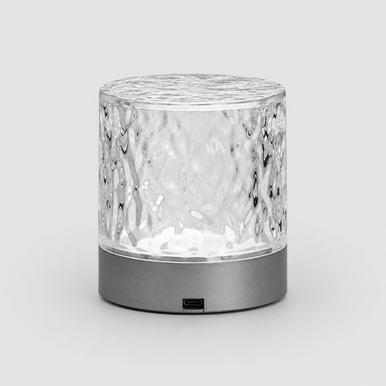 A modern cylindrical table lamp with a textured, hammered clear glass shade and a matte dark gray base, shown on a light gray background.