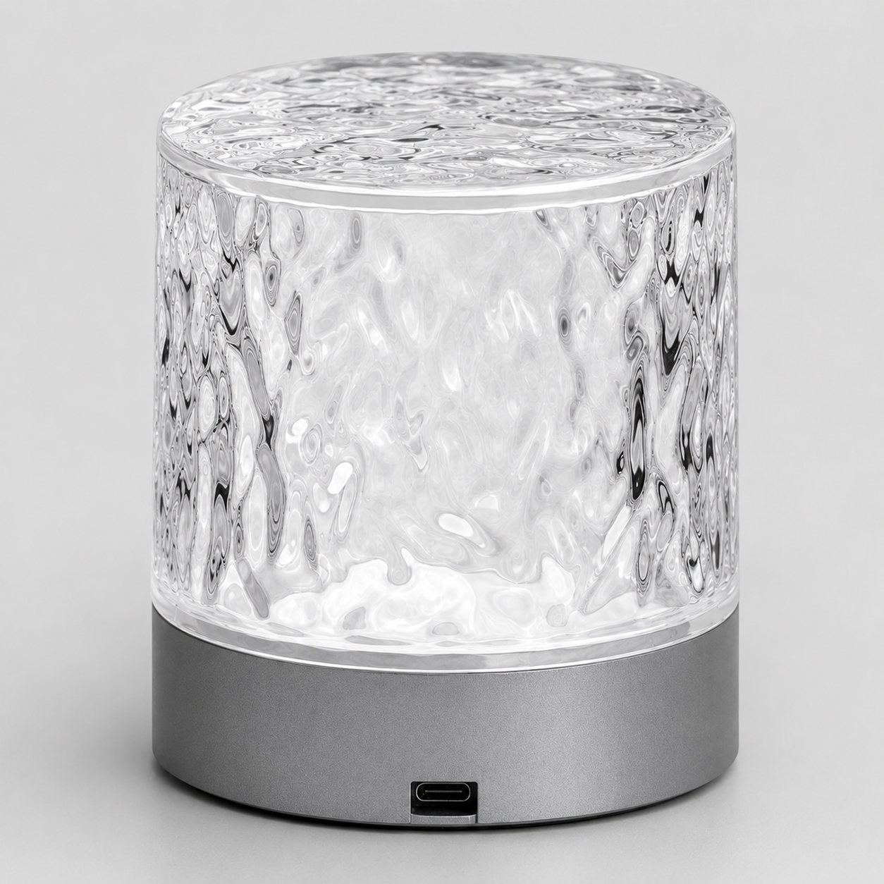 A modern cylindrical table lamp with a textured, hammered clear glass upper body and a matte dark gray metallic base, shown on a neutral light gray background.
