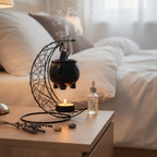 Black ceramic cauldron warmer above a lit tealight beside a bed.