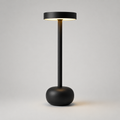 A modern black floor lamp with a wide circular shade, a slim central stem, and a rounded base, shown lit against a light gray studio background.