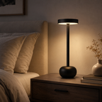 A modern black table lamp with a round base and flat circular shade, glowing warm white on a wooden nightstand next to a bed in a cozy bedroom setting.