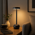 A modern black table lamp with a cylindrical shade and rounded base, glowing warm light on a wooden side table in a cozy living room setting.