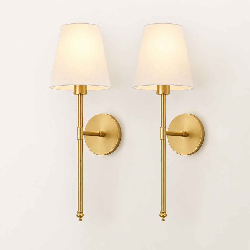 A pair of modern wall sconces with warm brass-finished metal arms and circular wall mounts, each holding a tapered off-white fabric shade that glows with a warm light, set against a plain off-white wall.