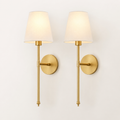 A pair of modern wall sconces with warm brass-finished metal arms and circular wall mounts, each holding a tapered off-white fabric shade that glows with a warm light, set against a plain off-white wall.