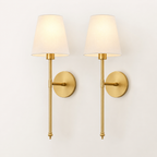 A pair of modern wall sconces with warm brass-finished metal arms and circular wall mounts, each holding a tapered off-white fabric shade that glows with a warm light, set against a plain off-white wall.