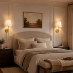 A beautifully styled bedroom featuring a bed dressed in neutral beige and cream linens, flanked by dark wood nightstands with lit wall sconces. A framed landscape painting hangs on the paneled wall above the bed, and a matching upholstered bench sits at the foot of the bed.