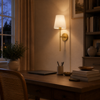 A warm brass wall sconce with a white fabric drum shade mounted on a beige wall above a wooden desk in a cozy home office setting.