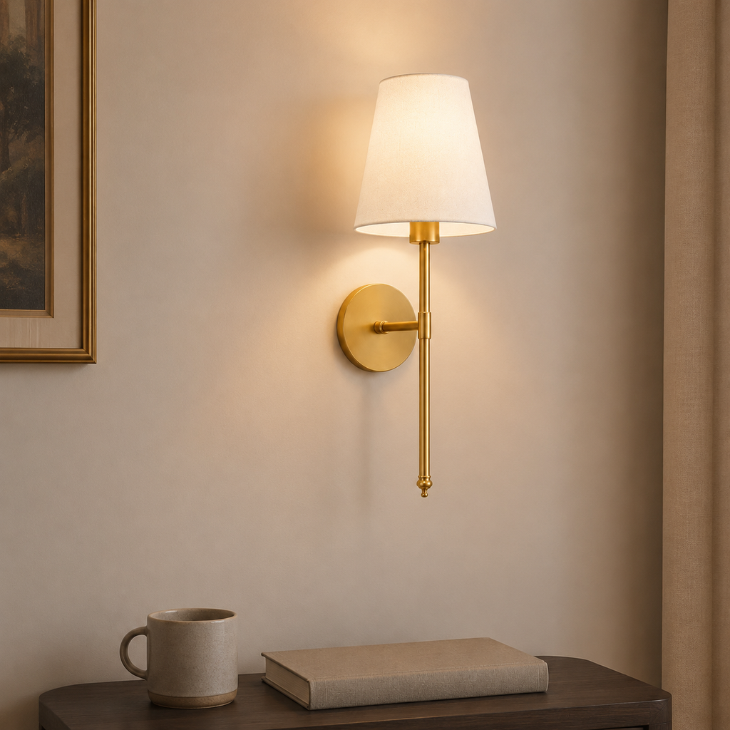 A modern wall-mounted sconce with a white tapered fabric shade and a brushed gold metal arm and circular backplate, shown lit on a beige wall above a dark wood table with a mug and book.