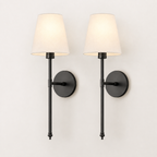 A pair of modern wall sconces with black metal fixtures and tapered off-white fabric shades, shown lit against a plain white wall.