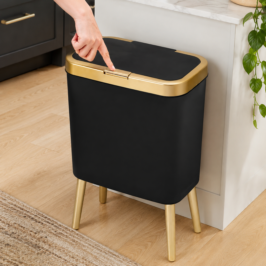 Black 15-liter trash can with gold trim beside kitchen cabinets as a hand opens the flip-top lid.