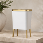 White trash can with elevated gold legs on a pale stone studio plinth.